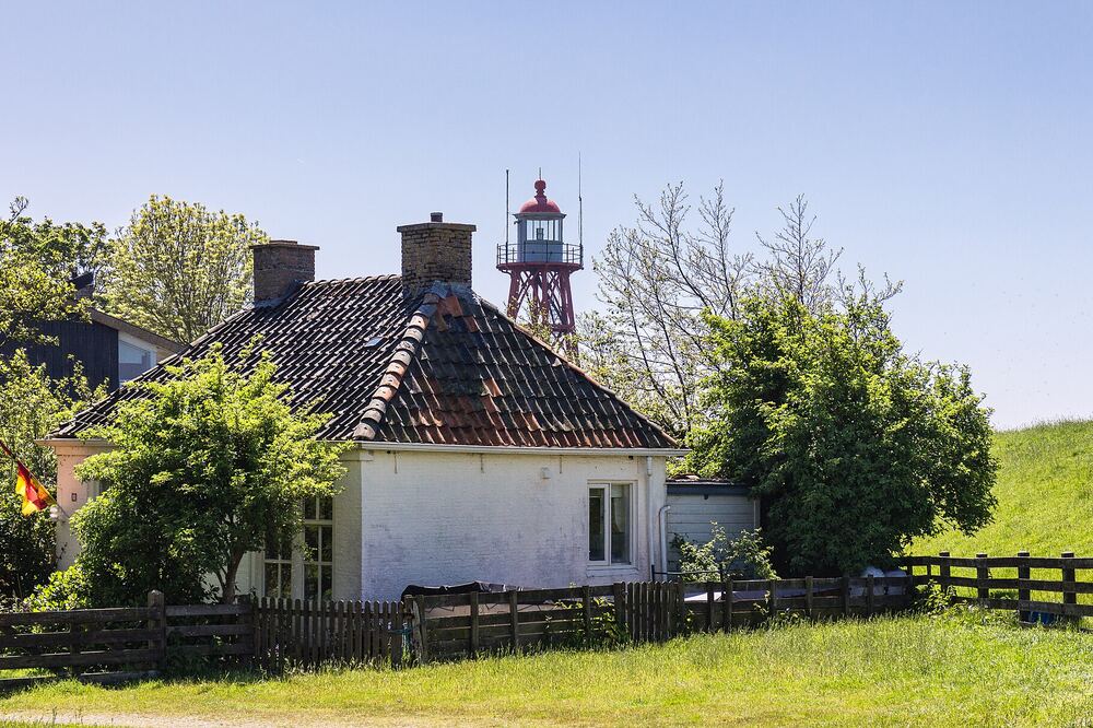 Wit huis met pannendak achter een houten hek, met daarachter een rood-witte ijzeren vuurtoren tussen de bomen
